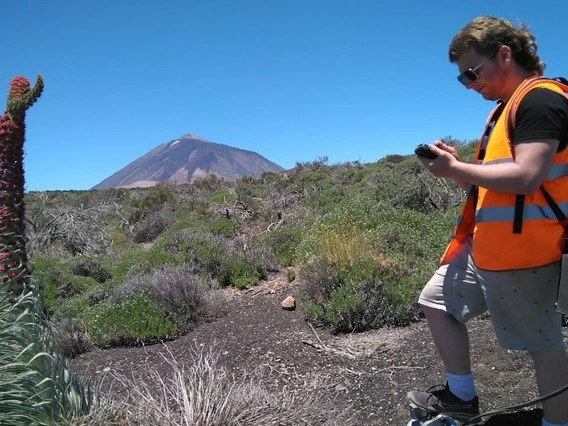 A young man in an orange vests stands in an area with volcanic rock.